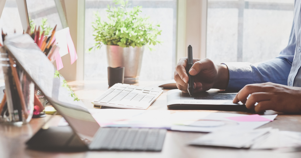 Hand holds electronic pen above tablet on top of a cluttered desk