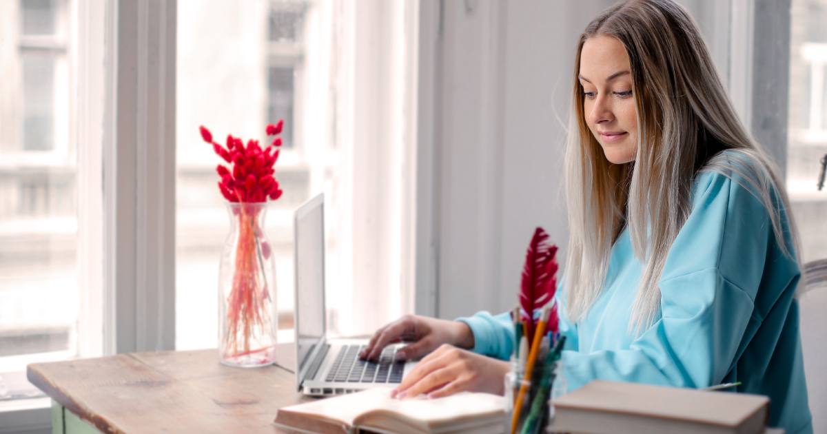 Person in blue sweater looks at book while hand rests on laptop