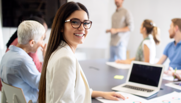 Person with glasses smiles at the camera while others talk during a meeting