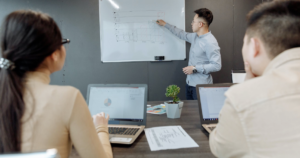 Person in front of whiteboard conducting sales goals meeting before people with laptops