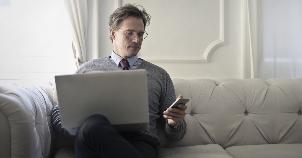Person in suit sits on couch while holding phone and balancing laptop on knee