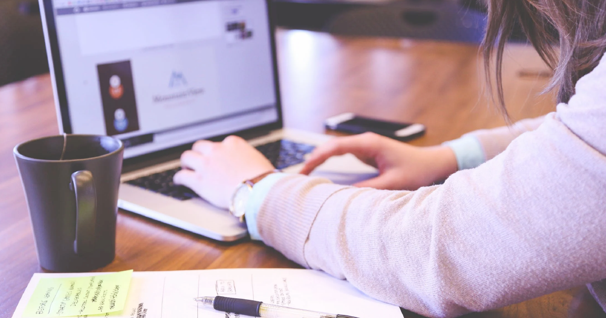 Person typing on computer on desk with cup and papers